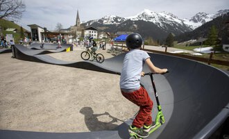 A skate park with children riding a scooter and a bike. In the background, mountains and a small town can be seen. | © Kleinwalsertal Tourismus | Frank Drechsel