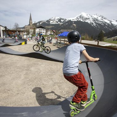 A skate park with children riding a scooter and a bike. In the background, mountains and a small town can be seen. | © Kleinwalsertal Tourismus | Frank Drechsel