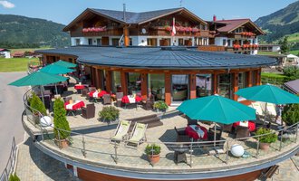 A modern hotel Birkenhöhe with a terrace, sun umbrellas, and cozy seating areas. In the background, green hills and mountainous landscapes are visible.