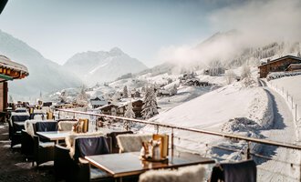 A cozy terrace with tables and chairs, surrounded by snow-covered mountains. The sky is clear and the landscape is picturesque.