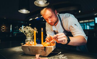 Our chef Felix Bantel artistically decorates a dish on a wooden board. On the table, there are candles and flowers that create a cozy atmosphere.
