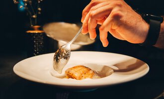 A hand is holding a spoon and serving a dish on a white plate. The background is dark, creating an elegant atmosphere.