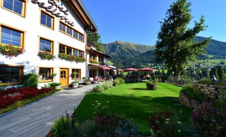 A beautifully designed house with balconies and blooming flowers. In the background, green mountains and a clear sky can be seen. | © Hotel Bellevue | I. Malouvier