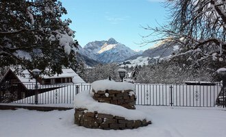 A winter landscape with snow-covered mountains in the background. The foreground shows a stone fireplace and snow-covered roofs. | © Hotel Bellevue | I. Malouvier