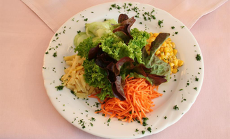 A colorful salad on a white plate with various types of vegetables. Underneath are visible green lettuce, carrot strips, corn, and cucumbers. | © Hotel Bellevue | Werbewind GmbH