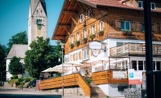 Ein traditionelles, holzverkleidetes Gebäude mit Balkon und Terrassen. Im Hintergrund ist ein Kirchturm und Bäume zu sehen.