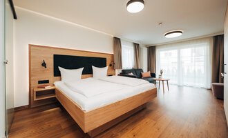 Ein modernes Hotelzimmer mit einem großen Doppelbett und Holzfußboden. Große Fenster lassen viel Tageslicht herein.