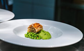A beautiful dish on a white plate, showcasing stuffed dumplings with a pea puree and fresh peas. The colors are vibrant and appealing. | © Verwöhn- & Wellnesshotel Walserhof| Frank Simon