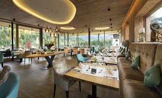 A modern restaurant with large windows and a view of nature. Stylish tables and cozy seating create an inviting atmosphere. | © Verwöhn- & Wellnesshotel Walserhof| R. Muehlbacher