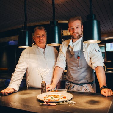 Two chefs stand in a modern kitchen behind a plate of elegantly arranged food. The atmosphere is inviting and creative, with soft lighting. | © Hotel Birkenhöhe | Stefan Klauser