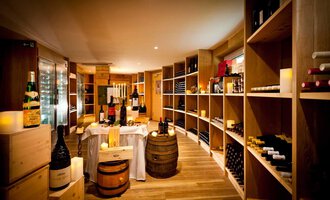 A cozy wine cellar with wooden shelves full of wine bottles. In the center, there is a table with candles and additional wines. | © Hotel Birkenhöhe | Fotograf Krug