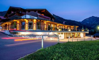 A modern building in alpine style, illuminated by warm light. In the background, impressive mountains are visible, while the sky glows at twilight. | © Hotel Birkenhöhe | GA Service Salzburg