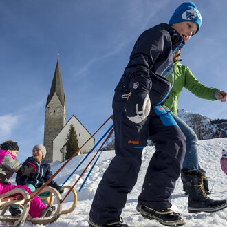 Children are sledding in the snow while adults in warm clothes accompany them. In the background, there is a church and the blue sky is visible. | © Kleinwalsertal Tourismus | Dominik Berchtold