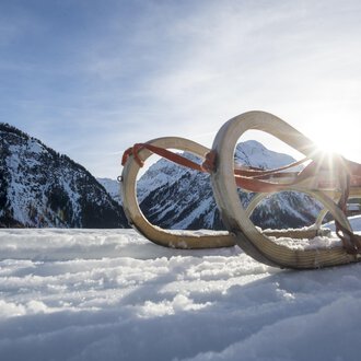 A sled lies in the snow, surrounded by mountains. The sun shines in the background, creating a wintry atmosphere. | © Kleinwalsertal Tourismus | Dominik Berchtold