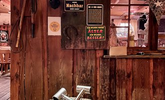 Ein rustikales Ambiente mit Holzinterieur und einem Baumstamm als Tisch. Auf dem Tisch liegen zwei Werkzeuge und an der Wand hängen verschiedene Schilder. | © s‘Älpele | Marco Mitterer