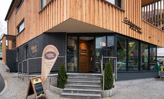 A modern restaurant with a wooden facade and large windows. The entrance area is designed to be welcoming, featuring a sign for the restaurant. | © Schönegger Käse-Alm