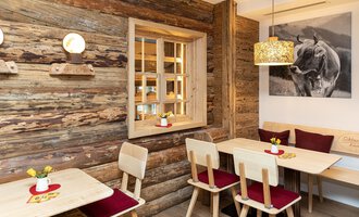 A cozy café with wooden-paneled walls and rustic furniture.  
Small flower vases are on the tables and a picture of an animal hangs on the wall. | © Schönegger Käse-Alm