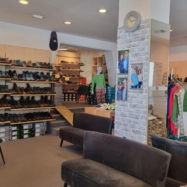 A shoe store with shelves full of shoes and a cozy seating area. Pictures hang on the walls and some clothing items are visible. | © Schuhhaus Schuster | Marietta Diehl