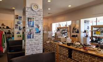 A shoe store with shelves full of shoes and clothing. On the left side, photos hang on the wall, and there is a large storefront that lets in plenty of light. | © Schuhhaus Schuster | Marietta Diehl