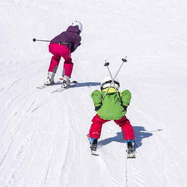 Two children skiing on a snow-covered slope. One child is wearing a green suit, the other a purple suit. | © Kleinwalsertal Tourismus | Dominik Berchtold