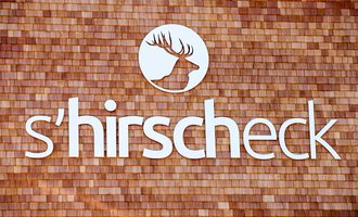 A stylish sign with the inscription "s'hirscheck". The background is made of wooden shingles and features a deer logo. | © Kleinwalsertal Tourismus | Frank Drechsel