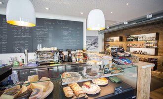 A cozy café with a selection of pastries and cheese. In the background, there is a board with the menu and shelves with drinks. | © Kleinwalsertal Tourismus | Frank Drechsel