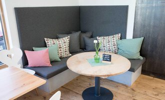 A cozy seating area with padded cushions in various colors. A round wooden table is in the center, decorated with a small plant. | © Kleinwalsertal Tourismus | Frank Drechsel