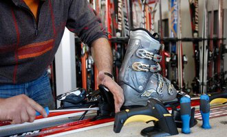A man is repairing ski equipment and assembling a ski boot. In the background, numerous skis are standing.