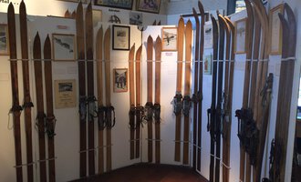 An exhibition featuring several wooden skis displayed on walls. In the background, photos and information about ski history are visible. | © Kleinwalsertal Tourismus | Blair Stevenson