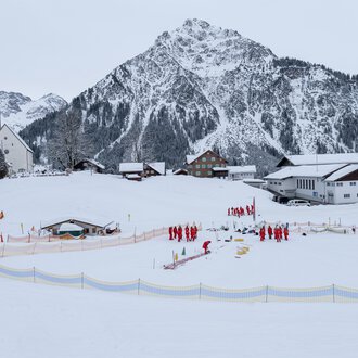 A snowy landscape with a village and a church in the foreground. In the background, steep mountains rise up, while a group of people in red jackets is active. | © Skischule Mittelberg | Karl Schuster