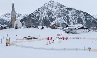 A snowy landscape with a village and a church in the foreground. In the background, steep mountains rise up, while a group of people in red jackets is active. | © Skischule Mittelberg | Karl Schuster