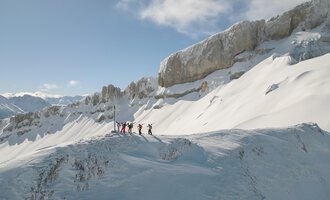 A group of people is standing on a snow-covered mountain peak. In the background, majestic rocks and a clear blue sky can be seen. | © Skischule Hirschegg GmbH