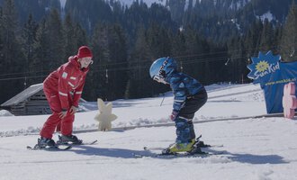 A ski instructor helps a child with skiing on a snow-covered slope. In the background, trees and a blue sign with the inscription "Ski-Kid" are visible. | © Skischule Hirschegg GmbH