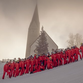 A group of people in red ski suits is standing in the snow in front of a church. The landscape is wintry and surrounded by fog. | © Skischule Mittelberg | Karl Schuster