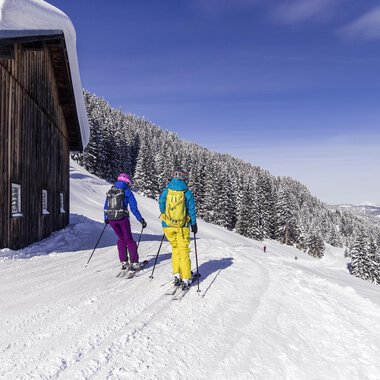 Zwei Skifahrer gehen einen verschneiten Weg entlang, vorbei an einer Holzhütte. Im Hintergrund sind schneebedeckte Berge und ein klarer blauer Himmel zu sehen. | © Kleinwalsertal Tourismus | Oliver Farys