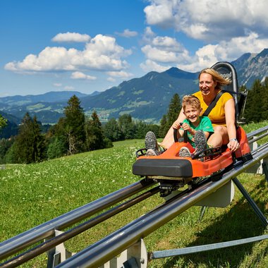A happy child and a woman are riding on a summer toboggan run. In the background, green mountains and a blue sky can be seen. | © OBERSTDORF · KLEINWALSERTAL BERGBAHNEN