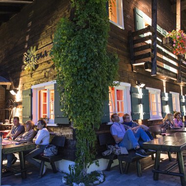 A cozy wooden house with colorful windows and plants. Several people are sitting at long tables on it, enjoying the evening atmosphere. | © Sonna Alp | Wolfram Riezler
