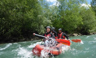 A group of people paddles on a red inflatable boat through fast currents. In the background, green trees and a blue sky can be seen. | © Spirits of Nature | Bernd Goller