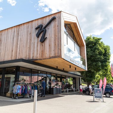 A modern shop with a wooden facade and large windows. In the foreground, clothing and bicycles are displayed, surrounded by a beautiful mountain landscape. | © Sport Kessler GmbH
