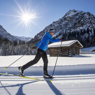 A cross-country skier in blue clothing is gliding through a snow-covered landscape. In the background, mountains and a small cabin can be seen under a clear blue sky. | © Kleinwalsertal Tourismus | Dominik Berchtold