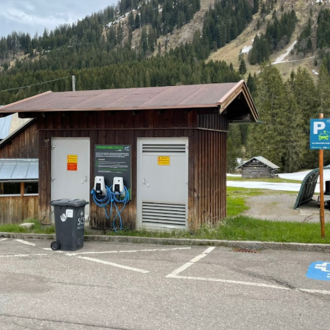 A small wooden building with charging stations for electric cars is located in an outdoor parking lot. In the background, green meadows and wooded hills can be seen. | © Kleinwalsertal Tourismus
