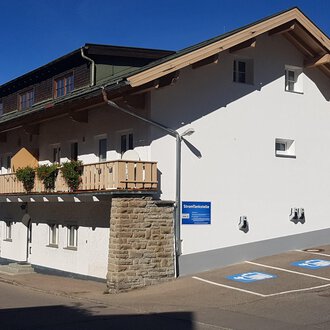 A modern building with a balcony and a parking space. The facade is bright and friendly, surrounded by mountains. | © Kleinwalsertal Tourismus
