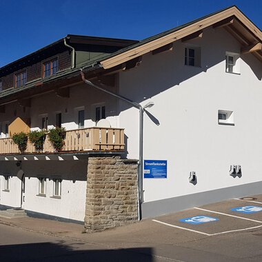 A modern building with a balcony and a parking space. The facade is bright and friendly, surrounded by mountains. | © Kleinwalsertal Tourismus