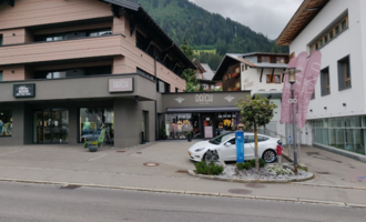 A modern shop in the mountains with a stylish facade. In front of the store, there is a white car and colorful flags are visible.