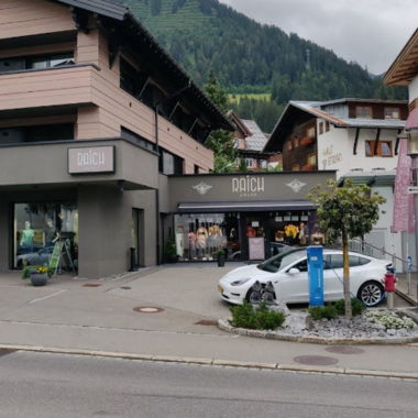 A modern shop in the mountains with a stylish facade. In front of the store, there is a white car and colorful flags are visible.
