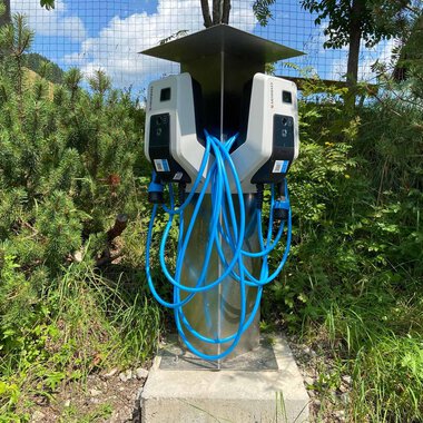 A modern charging station for electric vehicles with multiple charging ports. Surrounded by green vegetation and under a sunny sky. | © Kleinwalsertal Tourismus