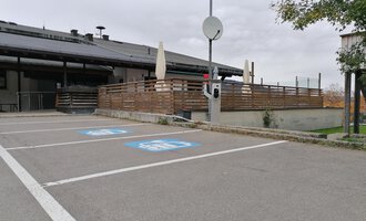 A parking lot with blue parking spaces for persons with disabilities. In the background, a building with a terrace and a gray sky is visible. | © Kleinwalsertal Tourismus