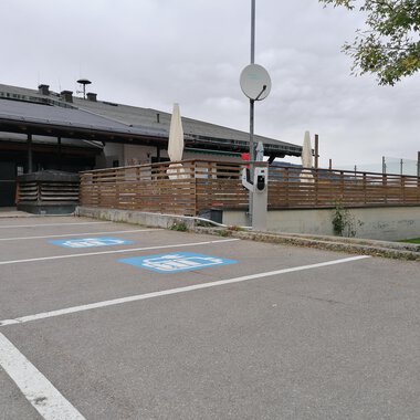 A parking lot with blue parking spaces for persons with disabilities. In the background, a building with a terrace and a gray sky is visible. | © Kleinwalsertal Tourismus