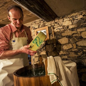 A man is standing at a wooden trough and pouring something in. In the background, stone walls can be seen. | © Kleinwalsertal Tourismus | Oliver Farys