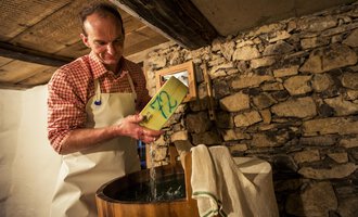 A man is standing at a wooden trough and pouring something in. In the background, stone walls can be seen. | © Kleinwalsertal Tourismus | Oliver Farys